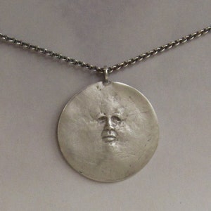 May include: A silver necklace with a round pendant featuring a textured face design.