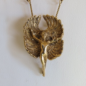 Puede incluir: Un collar con un colgante dorado que presenta una figura de querubín alado con los brazos extendidos. El querubín está representado de forma estilizada, casi abstracta, con plumas detalladas en las alas.