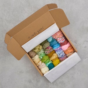 Op de afbeelding: Een kartonnen doos gevuld met kleurrijke garenbollen. De doos is open en de garenbollen zijn in rijen gerangschikt. Op de binnenkant van de doos staat "Thank You!" gedrukt.