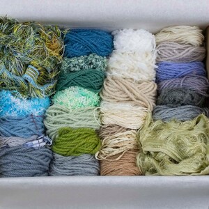 May include: A box filled with a variety of colourful yarn skeins and scraps. The yarn is in shades of blue, green, white, and brown.