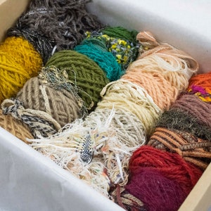 May include: A box filled with colourful yarn skeins in various textures, including a bright yellow, a deep green, a light peach, a brown with gold accents, and a deep red.