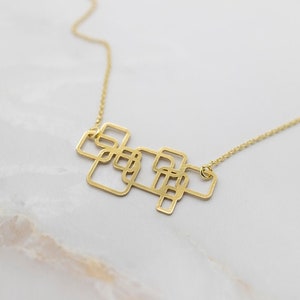 Puede incluir: Un collar dorado con un colgante geométrico compuesto por cuadrados entrelazados. La cadena es fina y el diseño es moderno y minimalista. El collar se muestra sobre una superficie de mármol blanco.