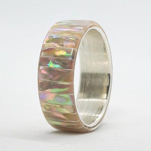 May include: A ring featuring an outer band inlaid with rectangular segments of iridescent abalone shell displaying pink, green, and gold hues. The interior of the band is made of a smooth, polished silver-toned metal with a reflective finish.