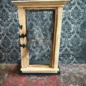 May include: A small wooden window frame with a cobweb inside. Three black bats are attached to the frame. The window is set against a dark patterned background.