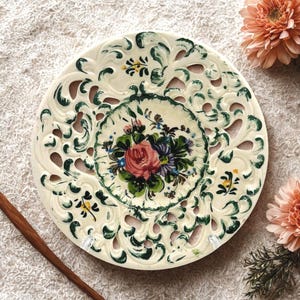 May include: Cream-colored decorative plate with a floral design. The plate features a central bouquet of pink, blue, and purple flowers, surrounded by green leaves. The rim has a cut-out design with green painted accents. A wooden stick is in the lower left corner.