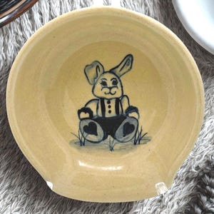 May include: A light yellow ceramic spoon rest with a blue and white hand-painted bunny design. The bunny is wearing overalls and sitting on grass. The spoon rest has a curved shape with a small handle.