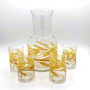May include: A clear glass pitcher and four matching glasses. The glassware features a wheat stalk design in shades of gold. The pitcher is taller than the glasses, and all pieces are set against a white background.