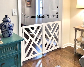 May include: White wooden pet gate with a geometric design, custom made to fit. The gate is open and is in a doorway with hardwood floors. The text "Custom Made To Fit!" is visible on the gate.