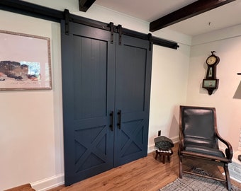 May include: A pair of dark blue barn doors with a black sliding track and black handles. The doors have a criss-cross design at the bottom. The doors are in a room with a white wall, a brown leather chair, and a fireplace.