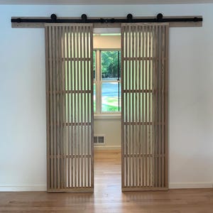 May include: Two wooden barn doors with a vertical slat design, mounted on a black metal track. The doors are open, revealing a window and the interior of a room. The doors are made of light-colored wood.