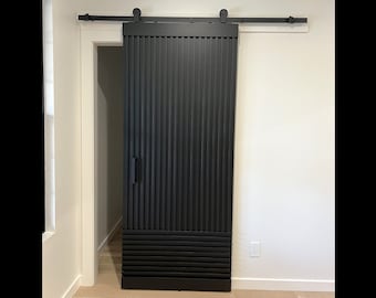 Op de afbeelding: Zwarte schuifdeur in schuur stijl met verticale houten latten en een horizontaal lattenpatroon aan de onderkant. De deur is gesloten en is gemonteerd op een zwart spoor.