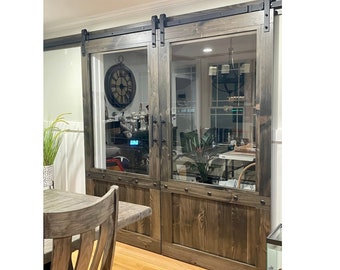 May include: A pair of rustic wooden barn doors with glass panels and black hardware. The doors are stained a dark brown color and have a distressed finish. The doors are open and reveal a room with a wooden table and chairs.