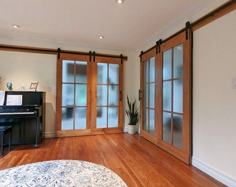 May include: Two sets of sliding barn doors with frosted glass panels and wooden frames. The doors are open, revealing a hardwood floor and a rug. A piano is in the background.