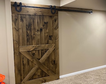May include: A rustic wooden barn door with a black metal track and hardware. The door has a large X-shaped design in the center and is stained a dark brown color.