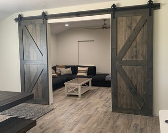 May include: Two dark brown wooden barn doors with black hardware and a diagonal wood design. The doors are open to reveal a living room with a sectional sofa and a coffee table.