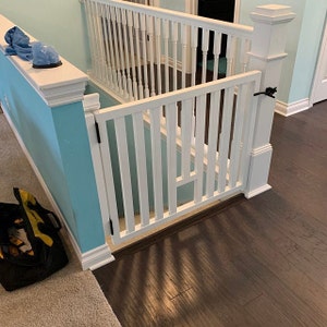 May include: White wooden baby gate with a black latch, installed at the top of a staircase. The gate is open and the stairs are visible in the background. The floor is dark wood.
