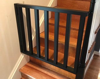 May include: Black metal baby gate installed at the top of a wooden staircase. The gate is open and has vertical bars.