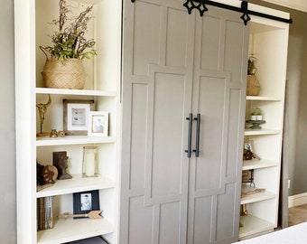 May include: White built-in bookshelf with sliding barn doors. The doors are gray with black hardware. The shelves are filled with decorative items, including plants, books, and framed photos.