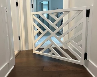May include: White wooden baby gate with a geometric design, featuring a diamond pattern. The gate is open and is in a hallway with hardwood floors.
