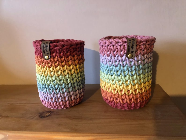 K&ouml;nnte beinhalten: Zwei handgefertigte H&auml;kel-Flaschenh&uuml;llen in einem Regenbogenverlauf. Die zylindrischen H&uuml;llen haben einen dunkelburgunderfarbenen oberen Teil und einen Regenbogen aus Farben, die in ein helles Rosa &uuml;bergehen. Jede hat ein Lederetikett mit der Aufschrift "Foliage & Flotsam".