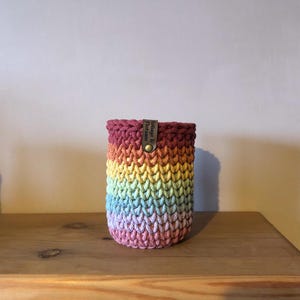 K&ouml;nnte beinhalten: Ein regenbogenfarbener H&auml;kel-Getr&auml;nkehalter mit einem Lederetikett mit der Aufschrift "Foliage & Flotsam". Der Halter ist zylindrisch und aus Garn in Rot-, Orange-, Gelb-, Gr&uuml;n-, Blau- und Lilat&ouml;nen gefertigt. Er steht auf einer Holzoberfl&auml;che.
