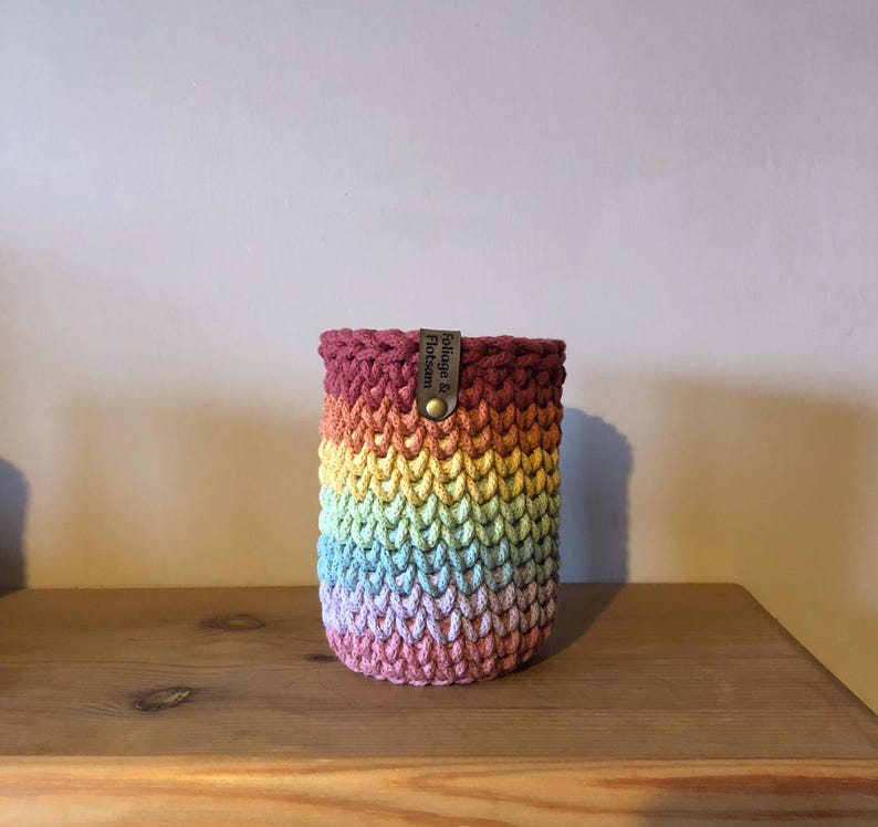 K&ouml;nnte beinhalten: Ein regenbogenfarbener H&auml;kel-Getr&auml;nkehalter mit einem Lederetikett mit der Aufschrift "Foliage & Flotsam". Der Halter ist zylindrisch und aus Garn in Rot-, Orange-, Gelb-, Gr&uuml;n-, Blau- und Lilat&ouml;nen gefertigt. Er steht auf einer Holzoberfl&auml;che.