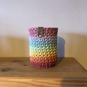K&ouml;nnte beinhalten: Ein handgefertigter, regenbogenfarbener H&auml;kelbecher. Der Becher hat eine zylindrische Form mit einem Farbverlauf von Lila nach Gelb. Ein kleines Lederetikett mit dem Text "Foliage & Flotsam" ist mit einem Messingknopf befestigt.