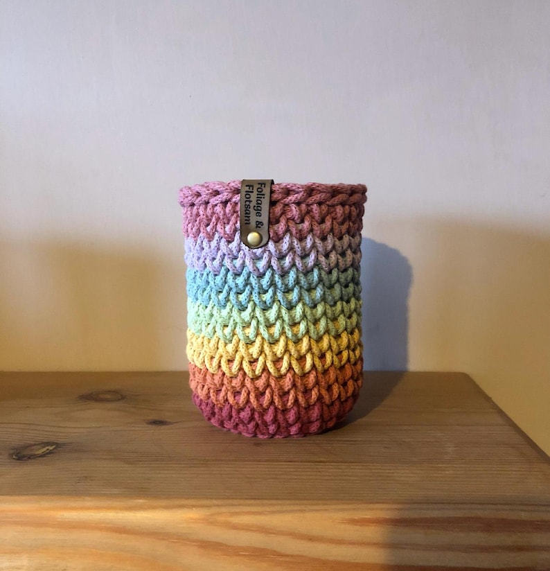 K&ouml;nnte beinhalten: Ein handgefertigter, regenbogenfarbener H&auml;kelbecher. Der Becher hat eine zylindrische Form mit einem Farbverlauf von Lila nach Gelb. Ein kleines Lederetikett mit dem Text "Foliage & Flotsam" ist mit einem Messingknopf befestigt.