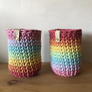 K&ouml;nnte beinhalten: Zwei Regenbogen-H&auml;kelk&ouml;rbe. Die K&ouml;rbe sind zylindrisch mit einem strukturierten Stichmuster. Die Farben gehen von Rosa &uuml;ber Blau, Gelb und Orange. Jeder Korb hat ein Lederetikett mit dem Text "Foliage Flotsam".