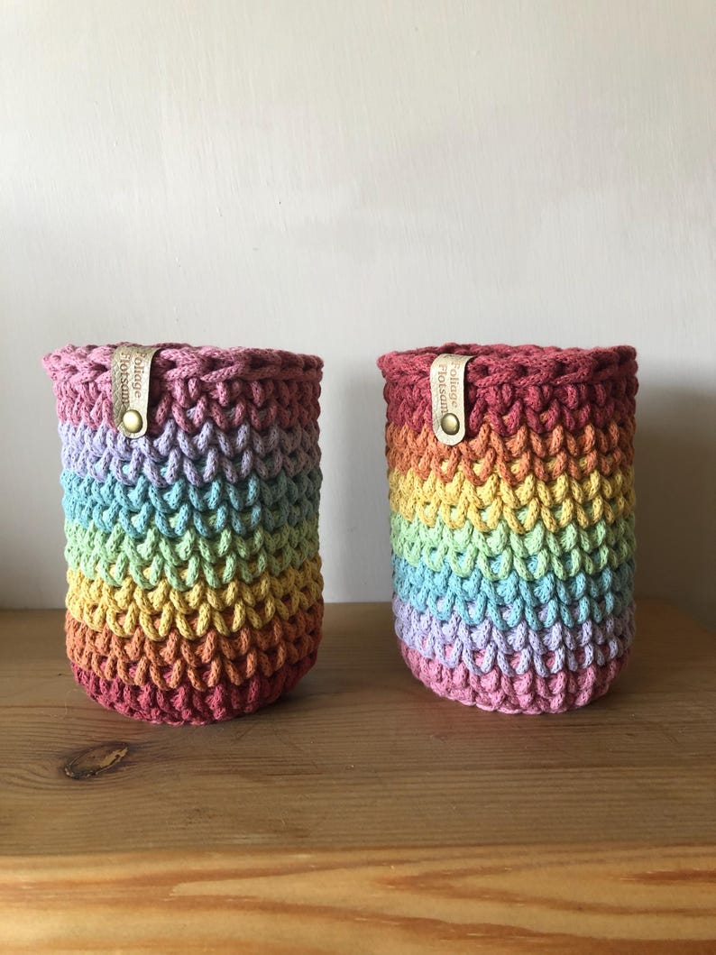 K&ouml;nnte beinhalten: Zwei Regenbogen-H&auml;kelk&ouml;rbe. Die K&ouml;rbe sind zylindrisch mit einem strukturierten Stichmuster. Die Farben gehen von Rosa &uuml;ber Blau, Gelb und Orange. Jeder Korb hat ein Lederetikett mit dem Text "Foliage Flotsam".