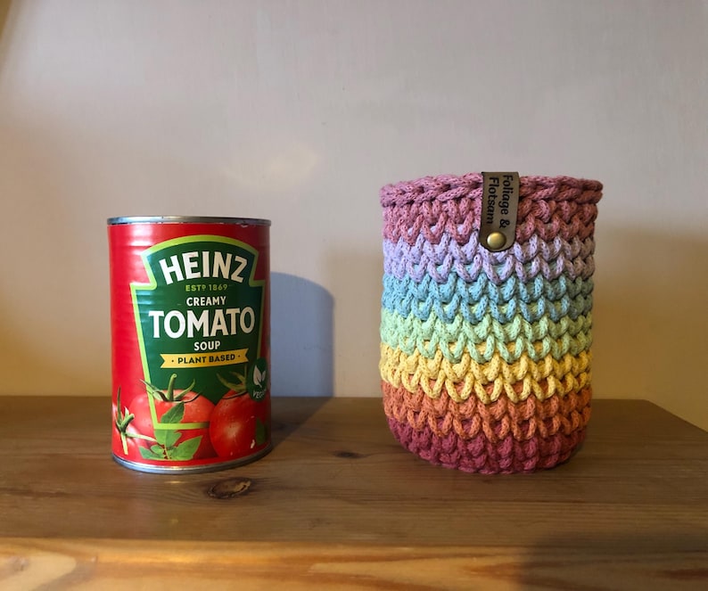 K&ouml;nnte beinhalten: Eine Dose Heinz Cremige Tomatensuppe neben einer Regenbogen-H&auml;kel-Getr&auml;nkeh&uuml;lle. Die Suppendose ist rot mit gr&uuml;nem und wei&szlig;em Text. Die H&uuml;lle ist zylindrisch, mit einem Lederetikett mit der Aufschrift "Foliage & Flotsam."