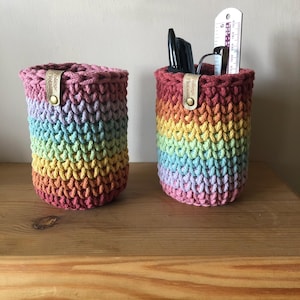 May include: Two rainbow-striped crochet pen pots. The pots are cylindrical with a textured stitch. Each has a leather tag with text and a metal snap. One pot contains pens, glasses, and a ruler. The pots sit on a wooden surface.