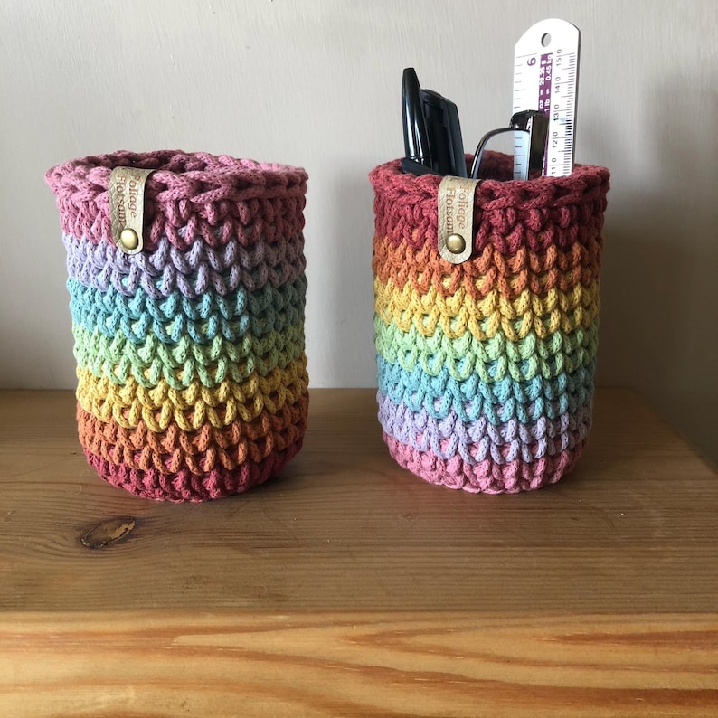K&ouml;nnte beinhalten: Zwei Regenbogen-H&auml;kelstiftehalter. Die Halter sind zylindrisch mit einem strukturierten Stichmuster. Jeder Halter hat ein Lederetikett mit Text und einen Metallschnappverschluss. Ein Halter enth&auml;lt Stifte, Brille und Lineal.