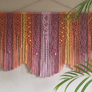 May include: A colourful macrame wall hanging in shades of yellow, orange, and purple. The handmade textile features intricate knotting patterns and a fringed bottom edge. The wall decor is suspended from a wooden dowel.