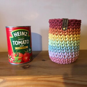 K&ouml;nnte beinhalten: Eine Dose Heinz Creamy Tomato Soup neben einer regenbogenfarbenen geh&auml;kelten Getr&auml;nkehalterung. Die Halterung hat ein Lederetikett mit der Aufschrift "Foliage & Flotsam". Die Suppendose ist rot mit gr&uuml;n-wei&szlig;em Text und Tomatenillustrationen.