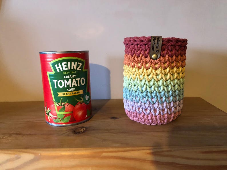 K&ouml;nnte beinhalten: Eine Dose Heinz Creamy Tomato Soup neben einer regenbogenfarbenen geh&auml;kelten Getr&auml;nkehalterung. Die Halterung hat ein Lederetikett mit der Aufschrift "Foliage & Flotsam". Die Suppendose ist rot mit gr&uuml;n-wei&szlig;em Text und Tomatenillustrationen.