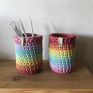 K&ouml;nnte beinhalten: Zwei Regenbogen-H&auml;kelhalter. Einer enth&auml;lt mehrere silberne H&auml;kelnadeln. Die Halter sind zylindrisch mit einem strukturierten Stichmuster. Die Farben gehen von Rosa &uuml;ber Gelb nach Blau. Jeder hat ein kleines Lederetikett mit Text.
