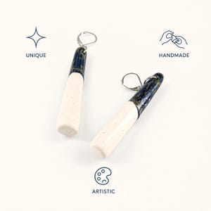 Puede incluir: Un par de pendientes colgantes con colgantes de cerámica cilíndricos y alargados. Cada pieza tiene una sección superior azul oscuro brillante con motas doradas y una sección inferior de color blanco roto mate. Incluyen ganchos de plata.