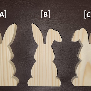 May include: Three wooden bunny cutouts in different designs. The bunnies are made of light-colored wood and have various ear shapes. The cutouts are displayed on a brown surface, perfect for Easter decorations.