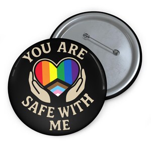 Puede incluir: Un botón negro con el texto "YOU ARE SAFE WITH ME" en blanco. El botón presenta un diseño de corazón arcoíris, con manos acunando el corazón. El botón es redondo y tiene un alfiler en la parte posterior.