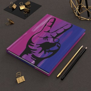Op de afbeelding: Een paars en blauw notitieboek met een zwart silhouet van een hand die het vredesteken maakt. Het notitieboek ligt op een grijs oppervlak met twee potloden.