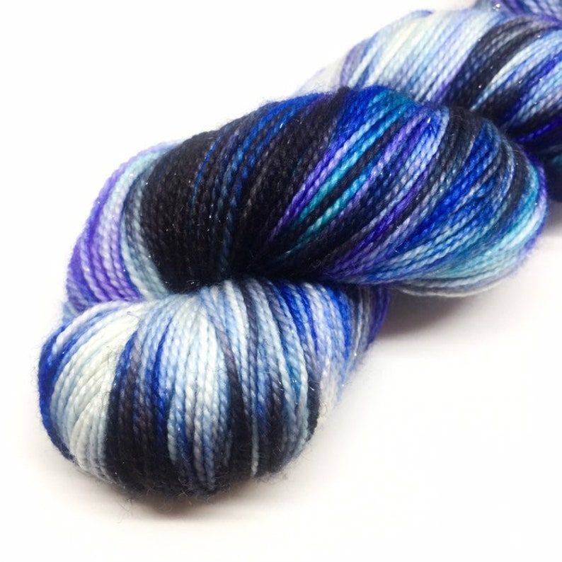 K&ouml;nnte beinhalten: Eine Garnrolle in einem blauen, violetten, wei&szlig;en und schwarzen Farbschema. Das Garn hat einen dezenten Glanz.