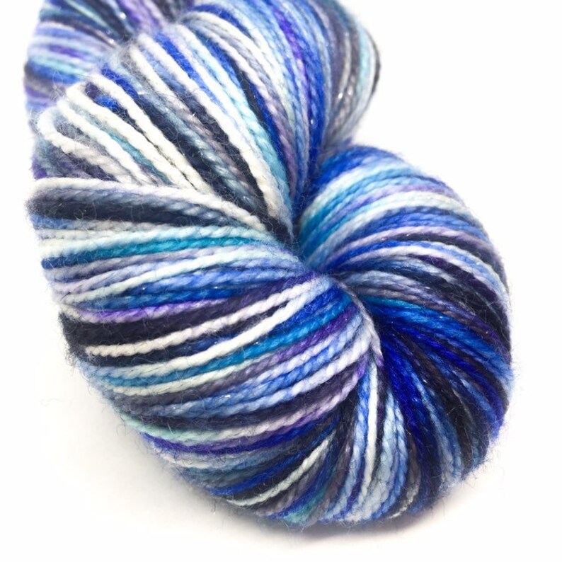 K&ouml;nnte beinhalten: Eine Garnrolle in einer blauen, wei&szlig;en und violetten Farbkombination mit einem dezenten Schimmer. Das Garn ist zu einer Kugel gewickelt und bereit zum Stricken oder H&auml;keln.