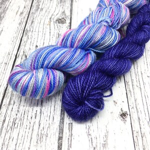 Könnte beinhalten: Zwei Knäuel Garn, eines ist ein mehrfarbiges blaues, rosa und weißes Garn, das andere ist ein einfarbiges dunkelviolettes Garn.