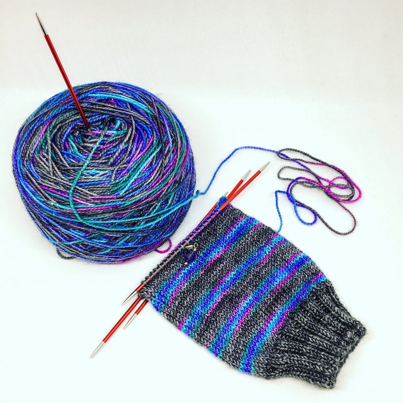 Puede incluir: Una bola de hilo de colores con una aguja de tejer roja que sobresale. Un calcet&iacute;n tejido parcialmente con rayas azules, moradas y grises est&aacute; sobre una superficie blanca con dos agujas de tejer.