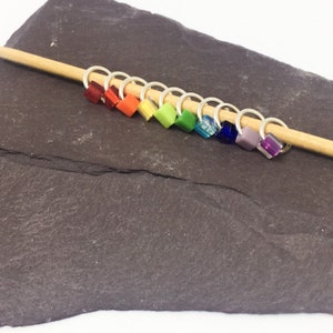 May include: A set of ten rainbow coloured glass bead stitch markers on a wooden knitting needle. The beads are square shaped and are arranged in a rainbow order from red to purple.