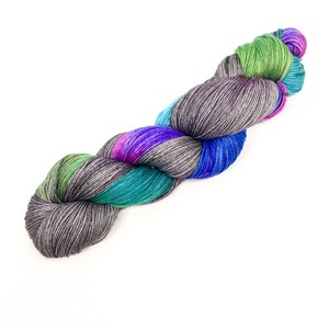 Puede incluir: Un ovillo de hilo con un patr&oacute;n de color variado. El hilo es una mezcla de gris, azul verdoso, verde, azul y morado.