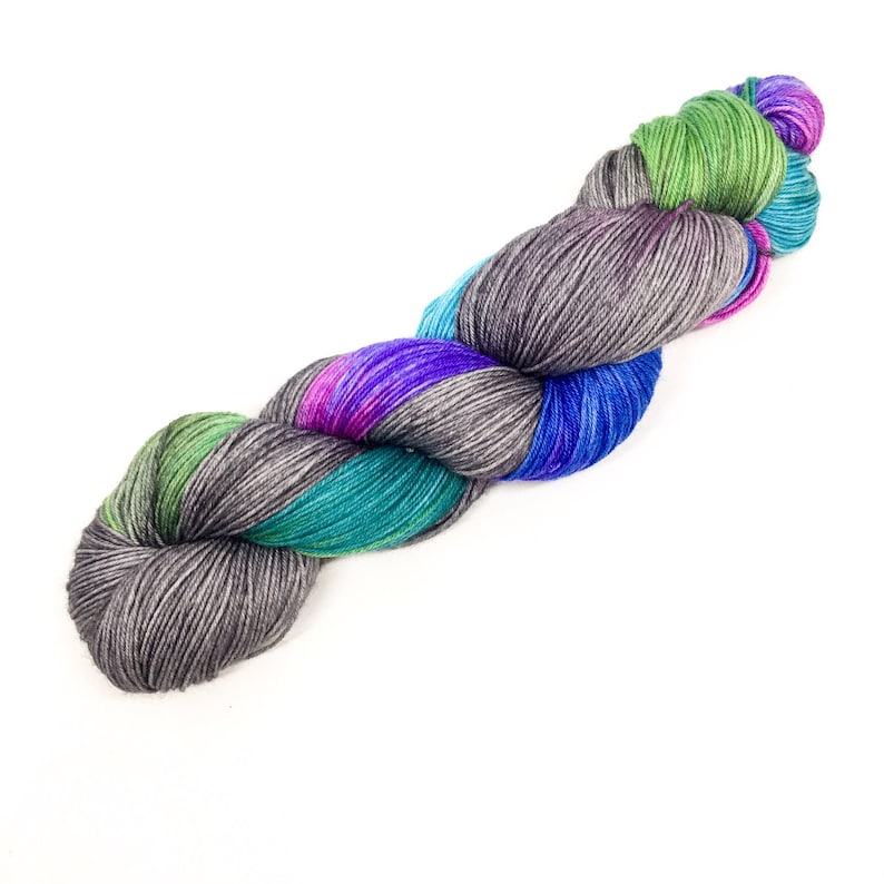 Puede incluir: Un ovillo de hilo con un patr&oacute;n de color variado. El hilo es una mezcla de gris, azul verdoso, verde, azul y morado.