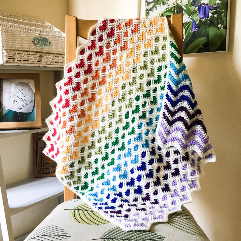 May include: A rainbow crochet blanket with a chevron pattern on the edge. The blanket is made with a variety of colors, including red, orange, yellow, green, blue, indigo, and violet.