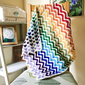 May include: A rainbow crochet baby blanket with a chevron pattern. The blanket is folded and draped over a wooden chair.
